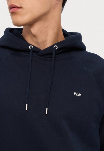 Tumši zilas krāsas kapuce, kas izgatavota no mīkstas auduma, ar sudraba krāsas caurumiem un auklām. Mazs balts izšūts logotips "W.W." uz krūtīm.
