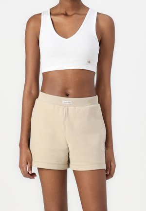 Frau trägt ein weißes bauchfreies Top und beige Calvin Klein-Shorts und steht vor einem einfarbigen weißen Hintergrund.