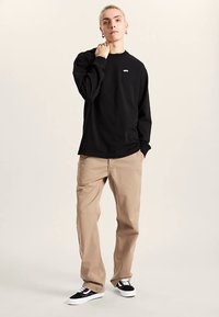 Schwarzes Langarmshirt mit kleinem Logo, beige Hose und schwarze Turnschuhe. Das Outfit zeichnet sich durch eine lockere Passform und weiche Stoffstruktur aus.