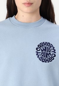 Hellblaues Sweatshirt mit geripptem Rundhalsausschnitt und einem auffälligen runden, navyfarbenen Grafik-Logo auf der Brust. Weicher Stoff.