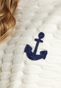 Valge kudumiga sviiter, millel on tekstureeritud muster ja tumehall broderi ankur emblem ees.