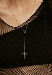 Collar de cadena de oro que presenta un largo cordón con un colgante en forma de cruz. La cruz tiene acentos decorativos y cuelga sobre una blusa de malla negra.
