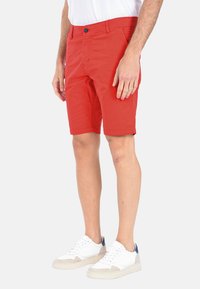 Shorts rossi in cotone con chiusura a bottone, tasche laterali e taglio dritto. Indossati con scarpe da ginnastica bianche con dettagli blu e beige.