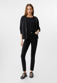 Mujer con sudadera negra con cremallera, camiseta gráfica negra, pantalones negros ajustados y sandalias negras con tachuelas, de pie con una mano en el bolsillo.