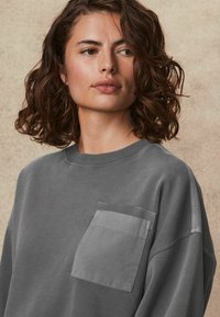 Grijze sweatshirt met een ronde hals, ribgebreide manchetten en een borstzak van contrasterende stof. Zachte textuur en casual ontwerp.