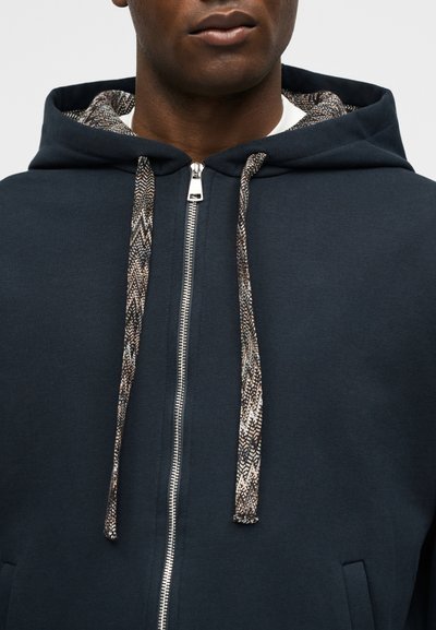 Άνδρας που φοράει σκούφο με φερμουάρ σε ναυτικό μπλε χρώμα με σχέδια στα κορδόνια και στη φόδρα της κουκούλας, ορατός από τους ώμους έως το στόμα.