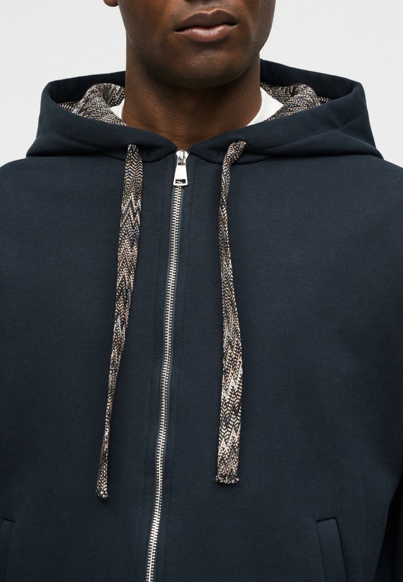 Homme portant un sweat à capuche bleu marine zippé avec des cordons à motifs et une doublure de capuche, visible des épaules jusqu'à la bouche.