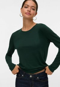 Frau in einem dunkelgrünen Langarmshirt und dunkelblauen Jeans, mit den Händen in den Bund gesteckt, vor einem schlichten hellen Hintergrund stehend.