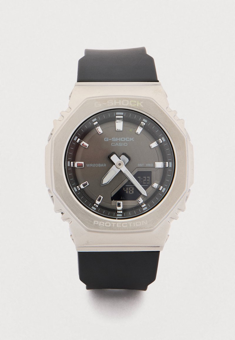 Ρολόι G-Shock με μεγάλο, στρογγυλό μεταλλικό κάσα, γκρι καντράν, μαύρο λουρί από καουτσούκ και φωσφορίζοντα δείκτες. Διαθέτει ψηφιακή οθόνη και αντοχή στο νερό.