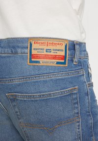 Modré džínové kalhoty z denimu s hnědou koženou štítkem s nápisem "Diesel Industry" a oranžovým prošíváním na kapsách.