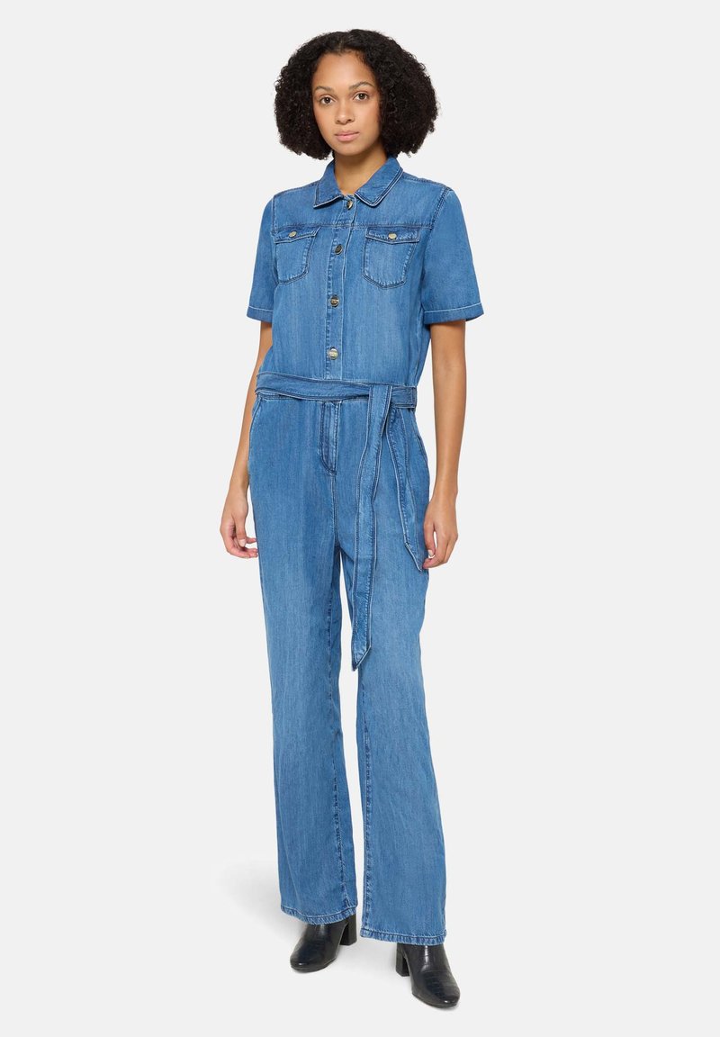 Combinaison en denim à manches courtes, devant à boutons, deux poches poitrine, ceinture en tissu, design à jambe large et couleur bleu clair.