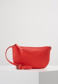 Červená kožená taštička s hladkým povrchem, se zipovým uzávěrem a odnímatelným náramkem na zápěstí. Obsahuje malou dekorační pásku na zipu.