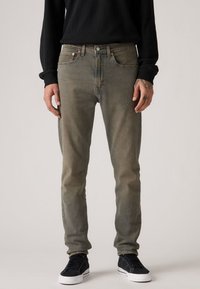 Slim-fit, faded green denim jeans with five pockets, copper hardware, and subtle wear. Paired with black sneakers, textured top.