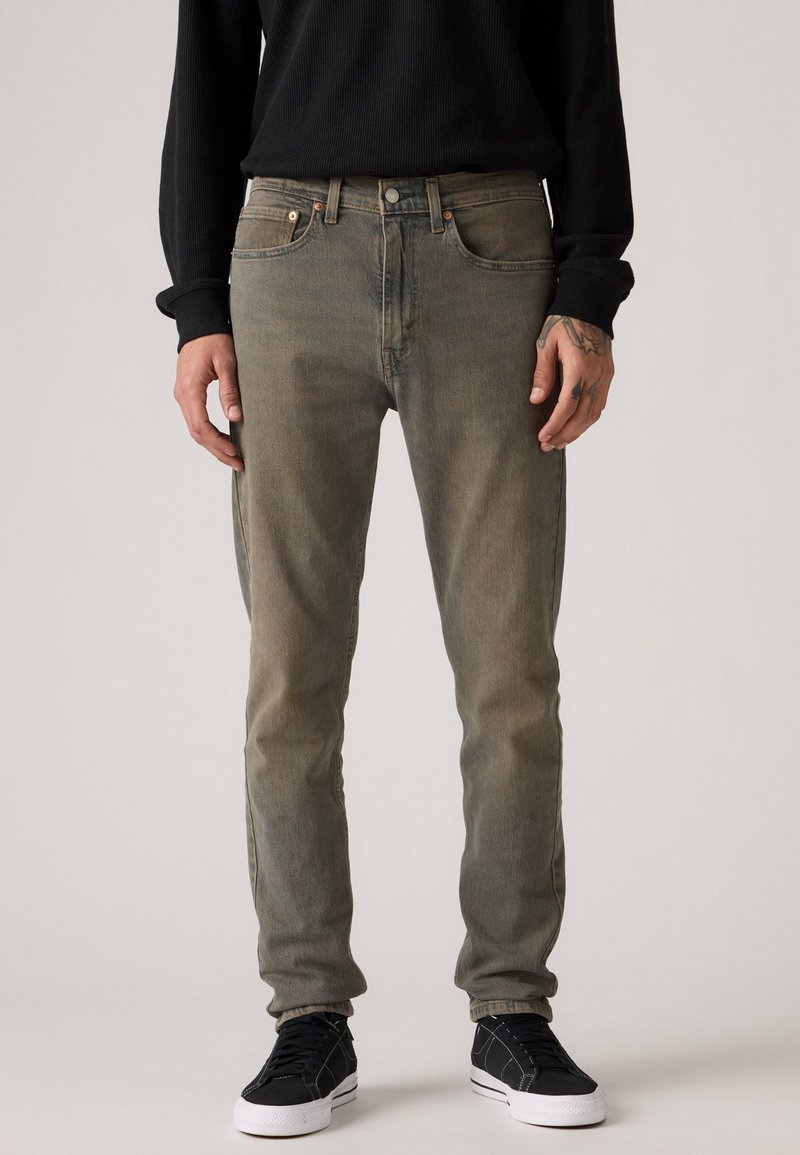 Slim-fit, faded green denim jeans with five pockets, copper hardware, and subtle wear. Paired with black sneakers, textured top.