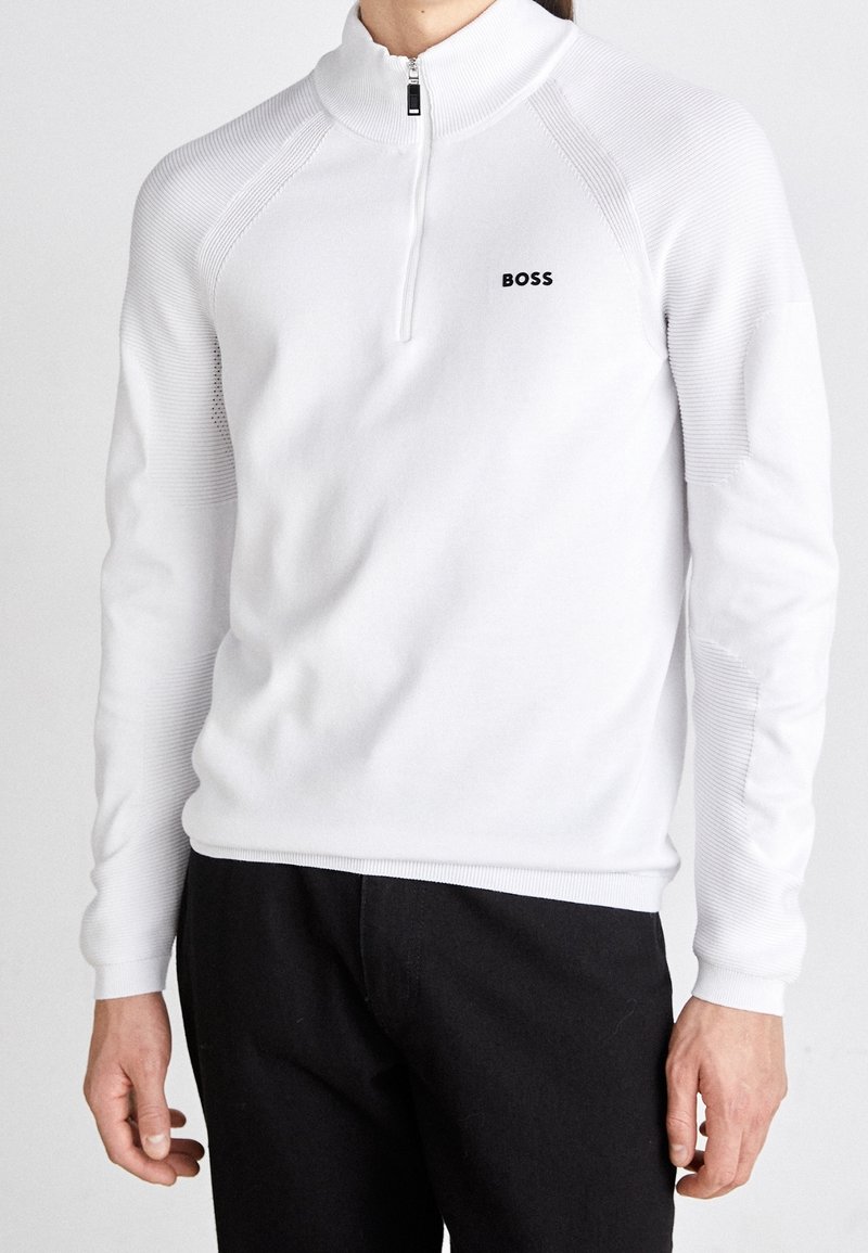 Homme portant un pull blanc à manches longues avec fermeture éclair au quart, logo noir « BOSS » sur la poitrine et pantalon noir sur fond uni.