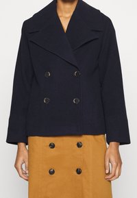 Manteau en laine bleu marine à double boutonnage avec de grands revers, doté de boutons noirs ronds et de manches trois-quarts, associé à une jupe beige boutonnée.