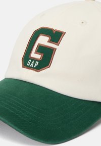 White cotton cap with a green bill, featuring a large green "G" and "GAP" in brown embroidery on the front. Textured fabric, structured design.