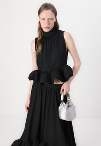 Robe noire sans manches avec un col haut et un ourlet froncé ; jupe noire avec taille froncée ; tenant un petit sac à main en satin argenté.