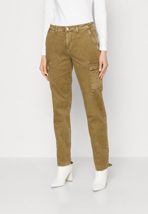 Kvinde iført brune cargobukser med sidelommer, hvid langærmet top og hvide ankelstøvler, stående foran en ensfarvet baggrund.