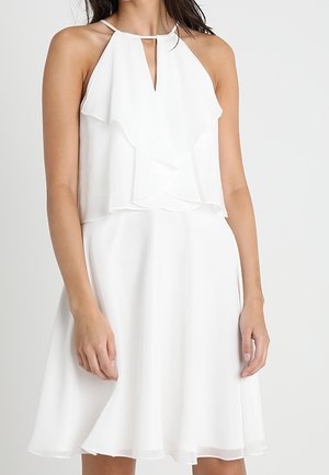 Kobieta ubrana w białą sukienkę bez rękawów z warstwowymi falbanami i dekoltem typu keyhole, stojąca na tle gładkim.