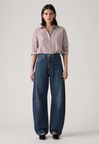 Chemise à rayures rouge et blanche avec des manches retroussées, associée à un jean en denim bleu foncé à jambes larges, avec des coutures contrastantes et des plis à l'avant.