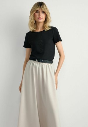 Kobieta o blond włosach do ramion, ubrana w czarną koszulkę z krótkim rękawem z cekinowym wzorem, beżową spódnicę i czarny pasek, stojąca na tle gładkiej ściany.