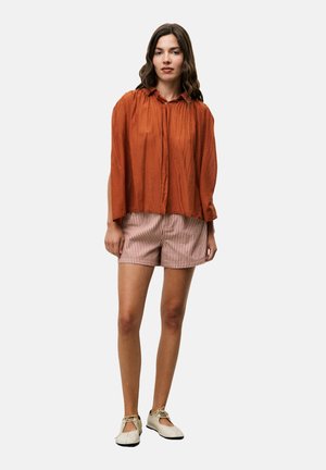 Roestkleurige, lichte blouse met een geplooide kraag, gecombineerd met lichtgestreepte shorts. Het model draagt beige schoenen met een ronde neus.
