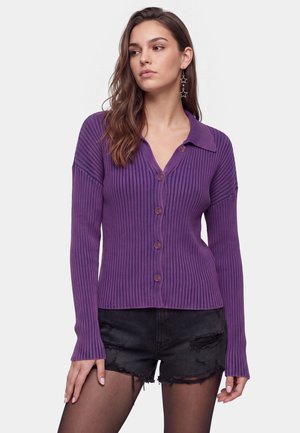 Young woman wearing a purple ribbed button-up sweater, black ripped shorts, and star-shaped dangling earrings, looking to the side.
