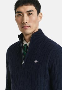 Marinefarbener Zip-Pullover aus strukturiertem Strickmaterial mit einem Zopfmuster. Verfügt über einen gerippten Kragen und ein dezentes Logo auf der Brust.