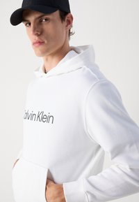 Sweatshirt alb cu glugă, având o buzunar frontal și logo-ul alb "Calvin Klein". Textură netedă cu manșete și tiv ribbate.