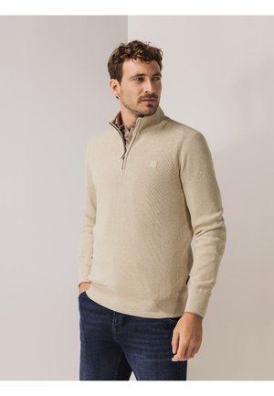 Beige, gestructureerde pullover met kwart-ritssluiting van geribd stof en een klein logo op de borst, gecombineerd met donkerblauwe jeans.