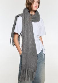 Grauer Wollschal mit fransigen Kanten, locker um den Hals drapiert. Weiche Textur zu einem schlichten weißen Hemd und hellblauen Jeans.