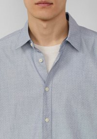 Himmelblaues Button-Up-Shirt mit einem subtilen Texturmuster. Verfügt über einen klassischen Kragen und cremefarbene Knöpfe, über einem weißen T-Shirt getragen.