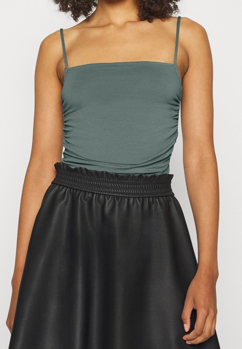Femme portant un débardeur turquoise plissé à encolure carrée avec de fines bretelles et une jupe en cuir noire taille haute avec une ceinture froncée.