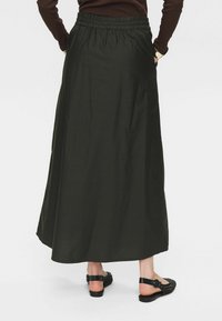 Jupe longue, vert foncé, en tissu doux avec une taille élastique. Présente une silhouette fluide et des poches latérales. Associée à des sandales noires.