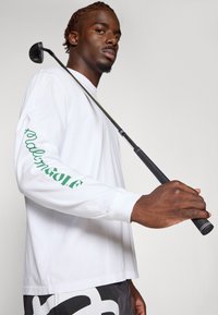 Camicia bianca a maniche lunghe con il testo verde "Palm Golf" sulla manica sinistra. Club da golf neri tenuti nella mano destra, modello che indossa pantaloni scuri.