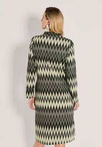 Frau mit blonden Haaren, die ein langärmliges, knielanges Kleid mit Zickzackmuster in Schwarz, Grau und Weiß trägt, mit dem Rücken zu einer schlichten Hintergrundwand.