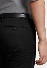 Muž v černých kalhotách s knoflíkovou zadní kapsou a černým pásem, oblečený se zakasanou kostkovanou košilí.