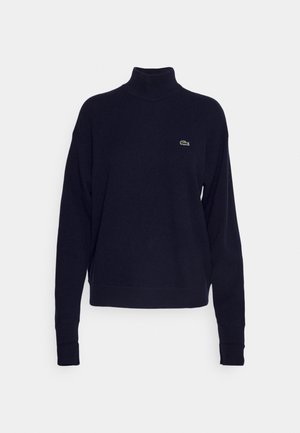 Navy blue turtleneck sweater made of soft fabric, featuring long sleeves and a small green logo on the chest. Simple, fitted design.