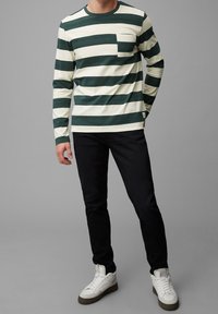 Mann in einem grün-weiß gestreiften Langarmshirt, schwarzen Hosen und weißen Sneakers vor einem grauen Hintergrund.