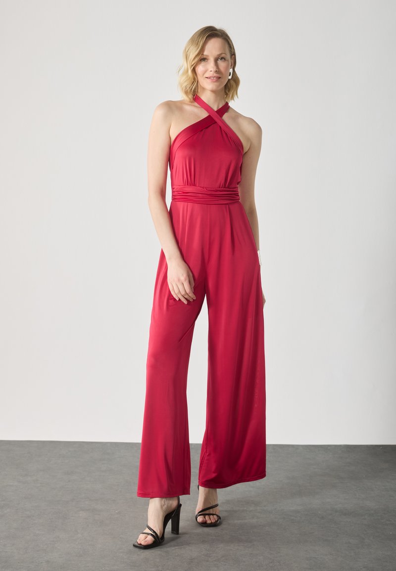 Rød ermeløs jumpsuit med halterneck-design, innsnørt midje og vide ben. Laget av et glatt stoff, kombinert med svarte høyhælte sandaler.