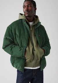Grønne corduroy jakke med hætte, der har elastiske manchetter og en afslappet pasform. Båret over en lysegrøn zip-up hættetrøje og jeans.