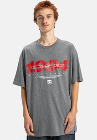 Jeune personne ayant les cheveux roux courts, portant un t-shirt gris oversized avec le numéro "1994" en rouge et le logo et le texte DC Shoe en blanc sur le devant.