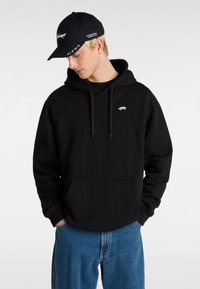 Felpa nera con cappuccio e tasca frontale, con un piccolo logo bianco, abbinata a jeans blu e un berretto nero con testo bianco e accenti di logo.