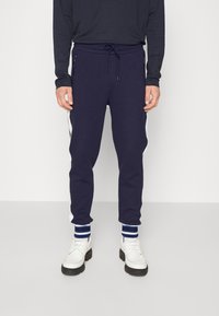 Marina joggare med dragsko i midjan, dragkedjefickor och vita sidostråkar. Kombinerade med vita skor och blårandiga muddar.