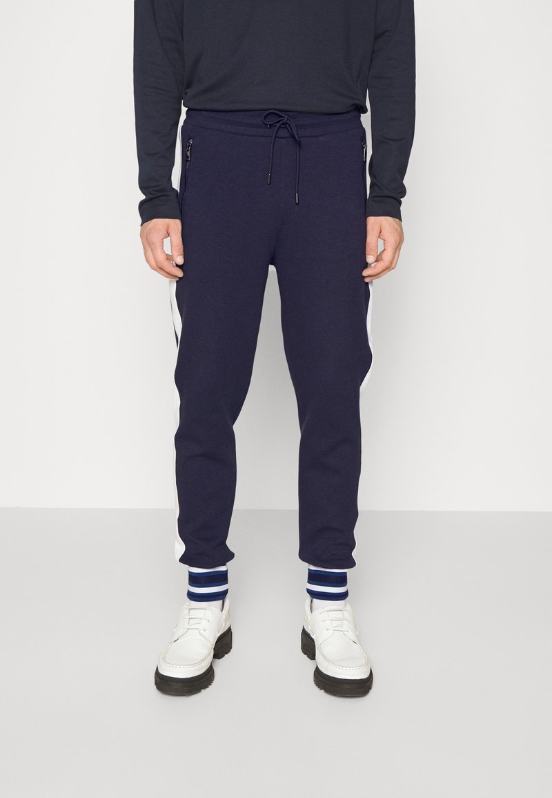 Marina joggare med dragsko i midjan, dragkedjefickor och vita sidostråkar. Kombinerade med vita skor och blårandiga muddar.