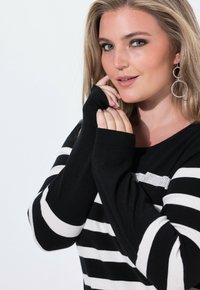Czarnobiała, pasiasta bluzka z długim rękawem o luźnym kroju, z otworami na kciuki i teksturowanym akcentem w okolicy dekoltu. Srebrne kolczyki w kształcie kół.