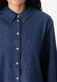 Denimskjorta i mörkblått med knapp-down krage, framficka och vita plastknappar. Har en texturerad tyg med subtila mönster.