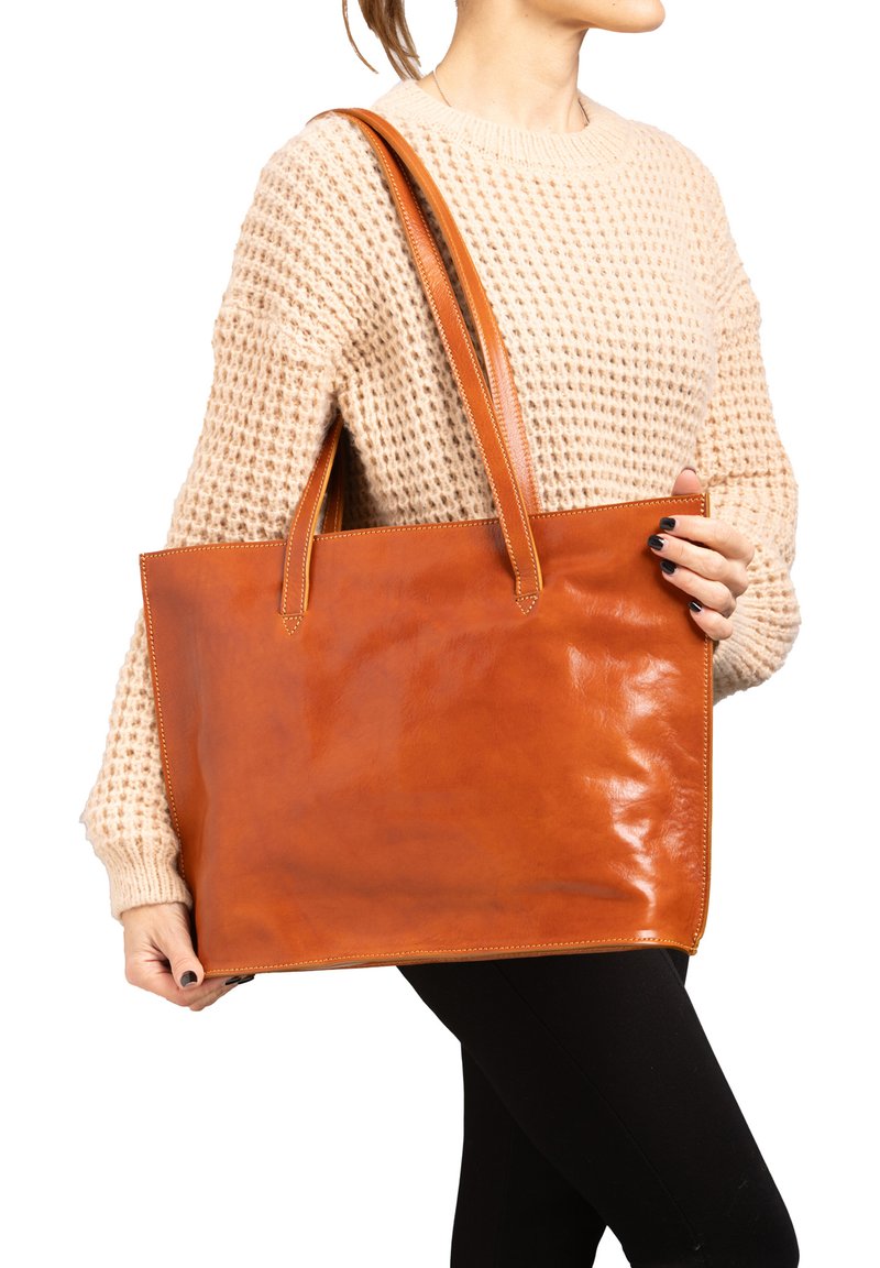 Tan leren totebag met een gladde textuur, twee schouderbanden en bovenste stiksels. Vlakke vorm met een open bovenkant, vastgehouden door een persoon in een beige sweater.