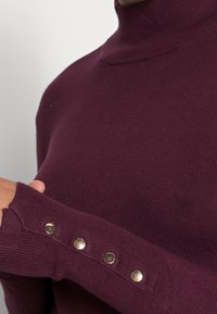 Bordowy golf z prążkowaną teksturą. Posiada złote guziki przy mankietach rękawów oraz dopasowany krój.